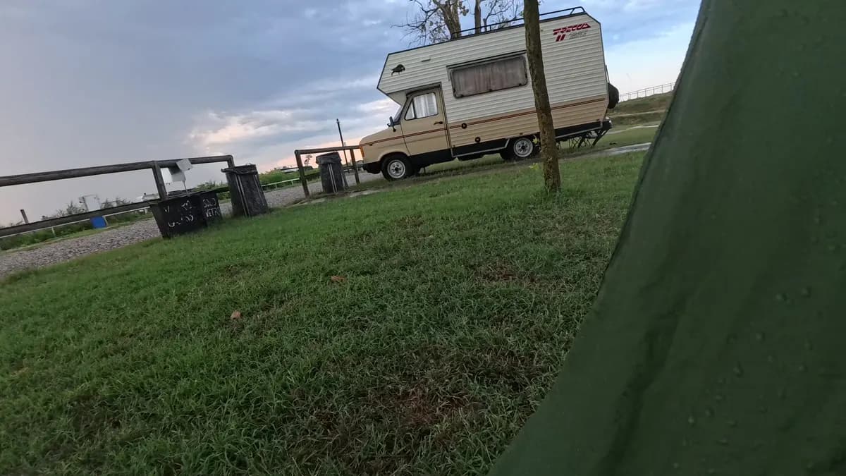 Blick aus dem Zelt auf die Wiese am Wohnmobilstellplatz Gorino, ein Holzzaun und ein altes Wohnmobil unter Gewitterwolken, August 2026.