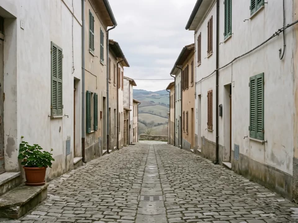 Eine gepflasterte Gasse zwischen verputzten Häusern mit Fensterläden, links ein Blumentopf auf einer Stufe, am Ende öffnet sich der Blick auf Hügel unter grauem Himmel
