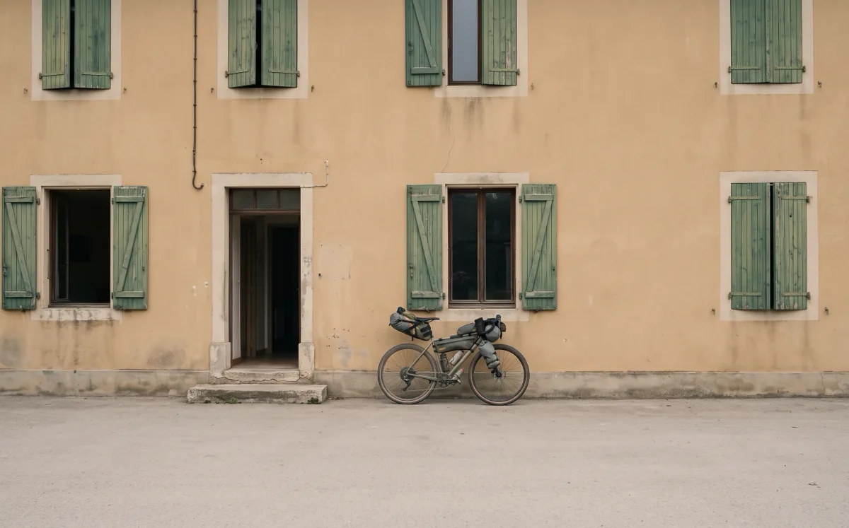 Die ockerfarbene Fassade eines Hauses mit grünen Fensterläden, die Haustür steht offen, rechts daneben lehnt ein bepacktes Gravelbike an der Wand, davor eine leere Straße