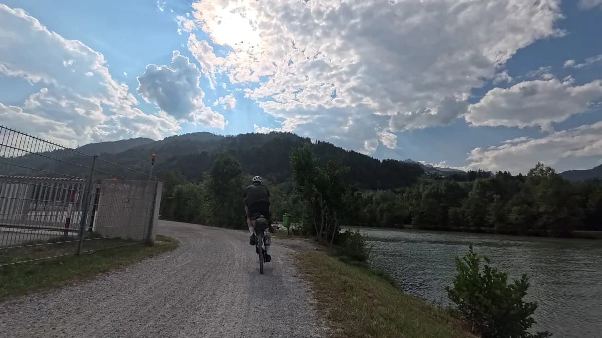 Schotterweg an der Salzach, ein Radfahrer mit Gepäck von hinten, darüber Wolken und Berge