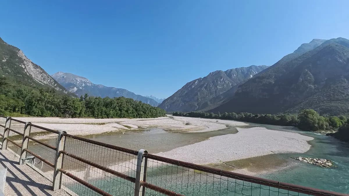 Breites, helles Kiesbett des Tagliamento zwischen bewaldeten Bergen, von einer Brücke mit Geländer aus gesehen