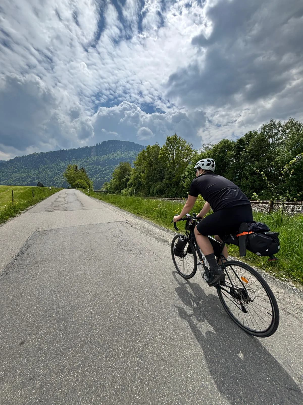 Person auf einem bepackten Rad mit Rennlenker, von hinten, auf einem schmalen Asphaltweg neben einer Bahnlinie, vorn bewaldete Berge unter hoch aufquellenden Wolken, 2025.